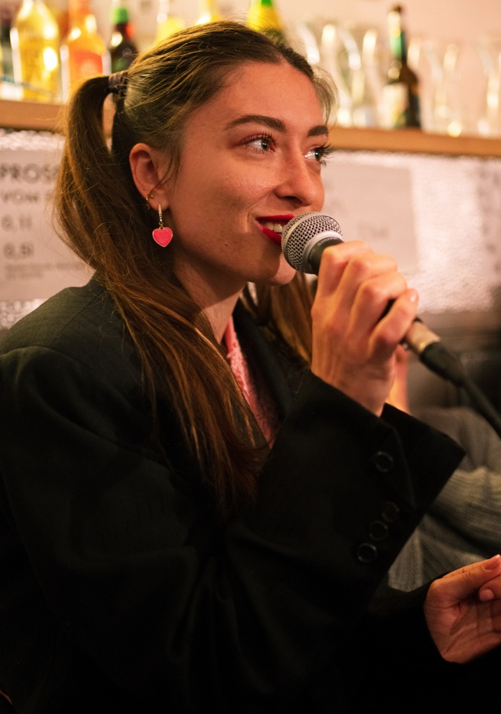 Claire Lefèvre has light brown hair tied back in pigtails, wears a heart shaped hearing, a pink top and black suit jacket, and is holding a microphone. Behind her is the blurred background of a bar, with many bottles and glasses